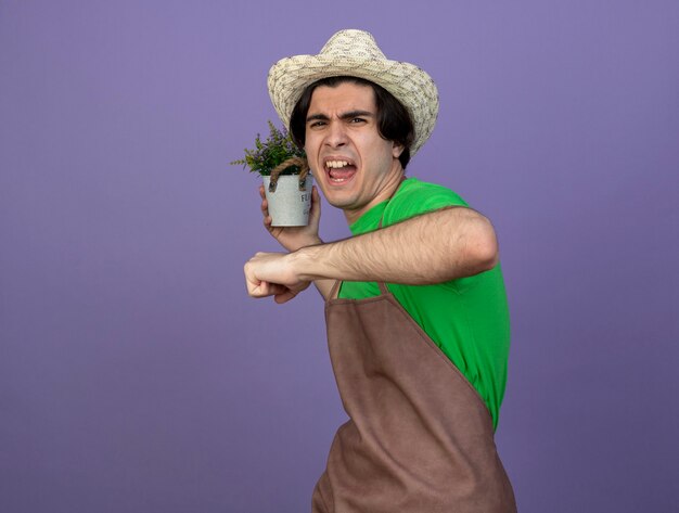 Angry young male gardener in uniform wearing gardening hat holding flower in flowerpot