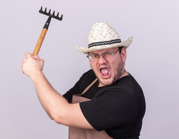 Angry young male gardener wearing gardening hat holding rake isolated on white wall