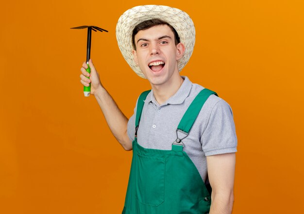 Angry young male gardener wearing gardening hat holds hoe rake