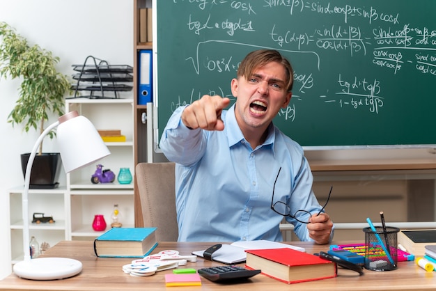 Angry young male teacher wearing glasses pointing with index finger  shouting with aggressive expression sitting at school desk with books and notes in front of blackboard in classroom