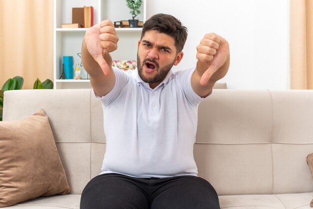 Angry young man in casual clothes looking  displeased showing thumbs down sitting on a couch in light living room