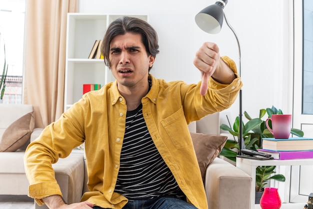 Free photo angry young man in casual clothes  showing thumbs down sitting on the chair in light living room