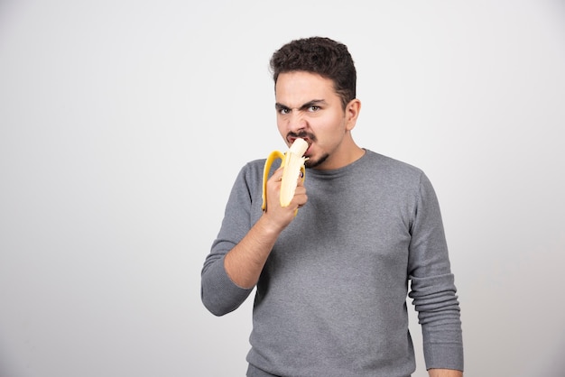 Free photo angry young man eating a banana over a white wall.