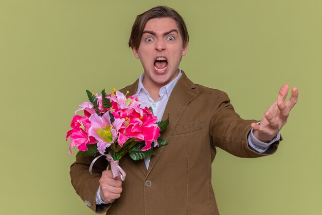 Angry young man holding bouquet of flowers looking at front yelling with aggressive expression with arm raised international women's day standing over green wall
