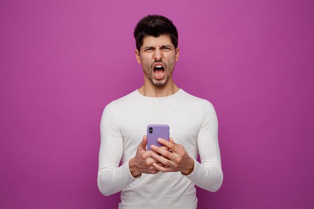 紫色の背景に分離されたカメラの叫びを見て携帯電話を保持している怒っている若い男