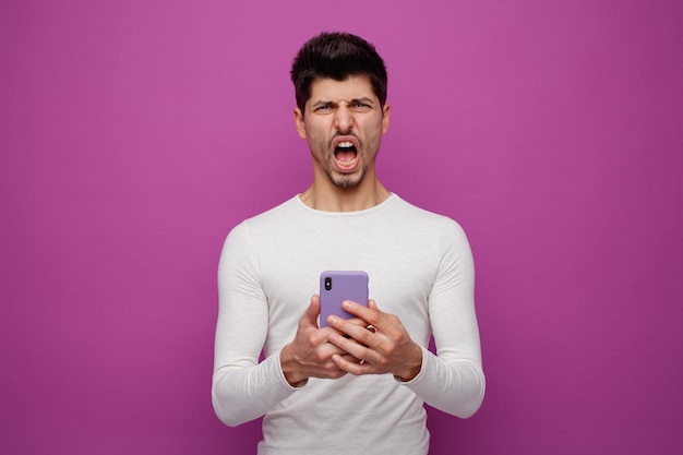 Злой молодой человек с мобильным телефоном смотрит в камеру и кричит на фиолетовом фоне