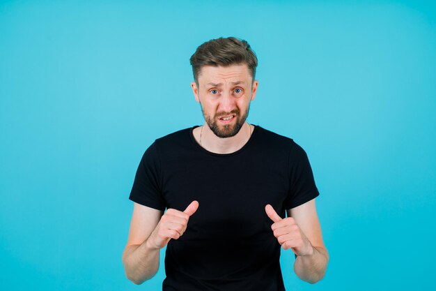 Angry young man is pointing himself with thumbs on blue background