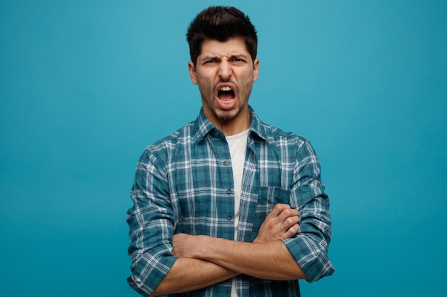 Angry young man looking at camera shouting with arms crossed isolated on blue background