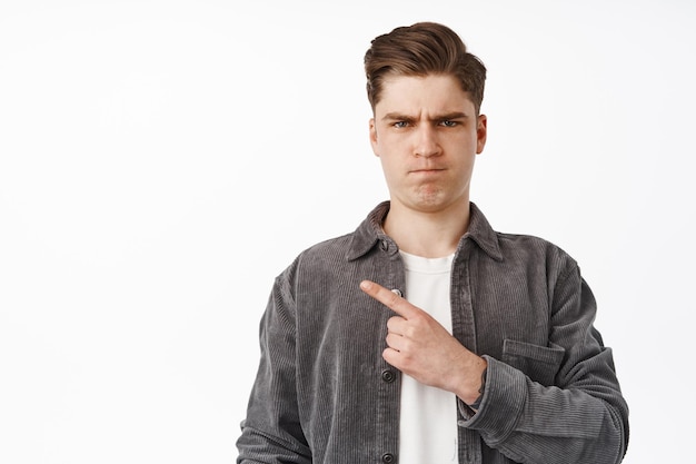 Angry young man pointing left, frowning and sulking, grimacing upset and complaining, showing something furious or outrageous, standing against white background