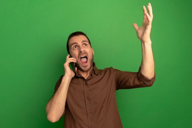 Foto gratuita giovane arrabbiato che parla sul telefono alzando la mano alzando lo sguardo isolato sulla parete verde