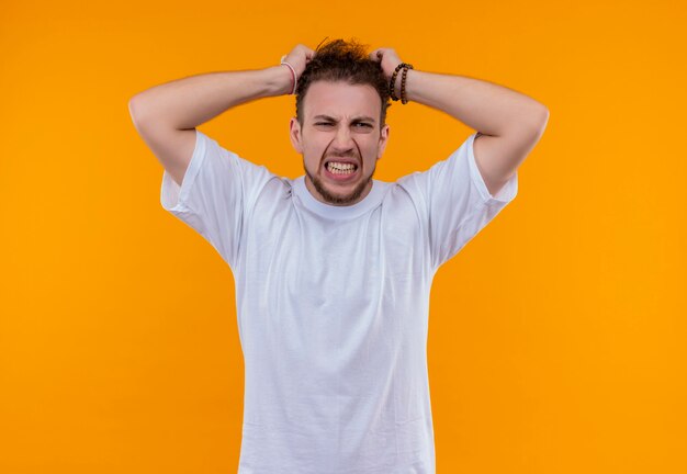 Angry young man wearing white t-shirt grabbed head on isolated orange wall