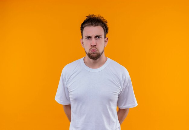 Angry young man wearing white t-shirt put his hand on back on isolated orange wall