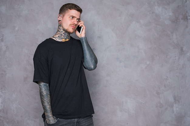 Free photo an angry young man with tattooed on his hand talking on mobile phone against grey wall