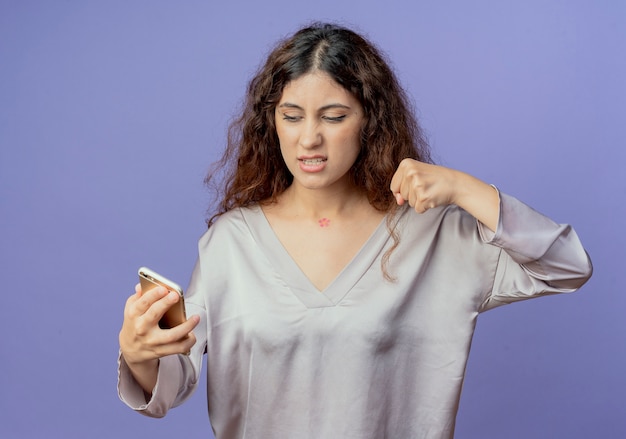 Angry young pretty girl holding and looking at phone and raising fist 