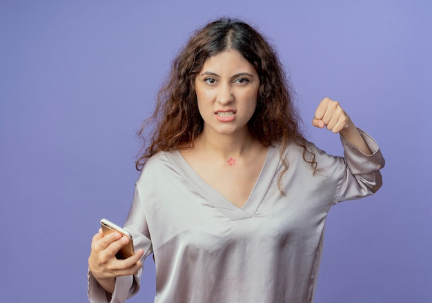 Free photo angry young pretty girl holding phone and raising fist isolated on blue wall