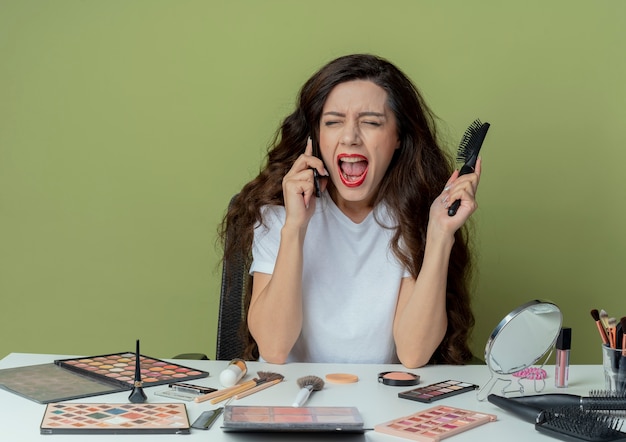 Arrabbiato giovane bella ragazza seduta al tavolo per il trucco con strumenti di trucco che tiene pettine e parlando al telefono urlando con gli occhi chiusi isolati su sfondo verde oliva