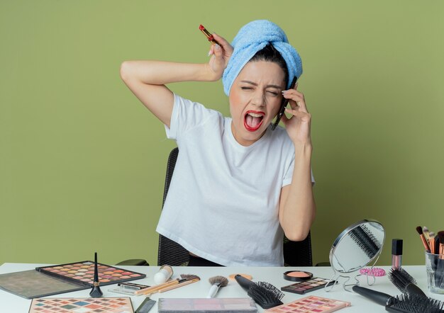 Foto gratuita arrabbiato giovane bella ragazza seduta al tavolo da trucco con strumenti per il trucco e con l'asciugamano sulla testa parlando al telefono toccando la testa con il rossetto in mano urlando con gli occhi chiusi sullo spazio verde oliva