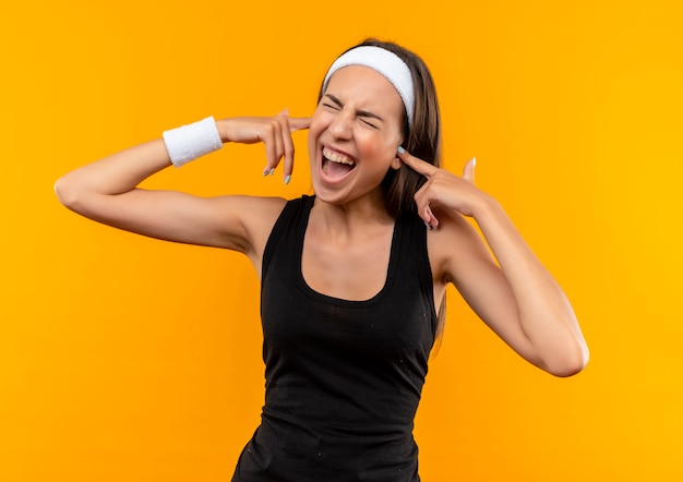 Angry young pretty sporty girl wearing headband and wristband putting fingers in ears with closed eyes isolated on orange wall