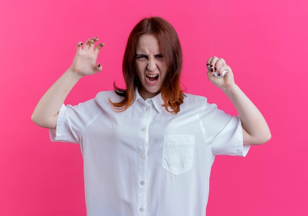 Angry young redhead girl crushing paper and doing tiger style isolated on pink wall