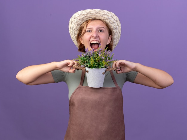 コピースペースと紫色の壁に分離された植木鉢に花を保持しているガーデニング帽子をかぶって怒っている若いスラブ女性庭師