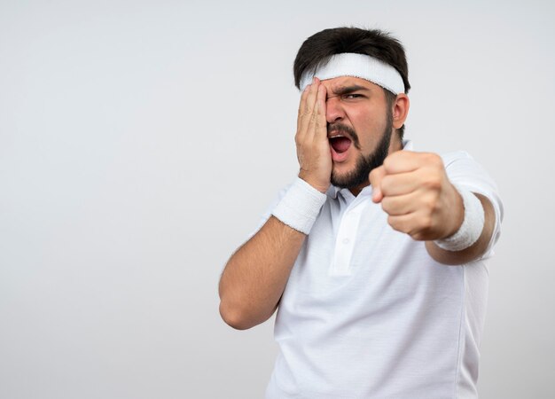 Сердитый молодой спортивный человек с повязкой на голову и браслетом, протягивающим кулак, положив руку на лицо, изолированное на белой стене с копией пространства