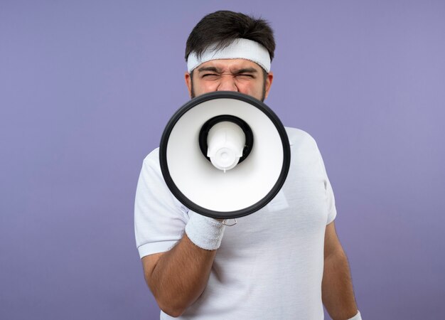 Angry young sporty man wearing headband and wristband speaks on loudspeaker isolated on green wall