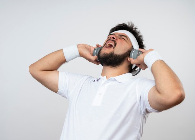 Angry young sporty man with closed eyes wearing headband and wristband with headphones isolated on white