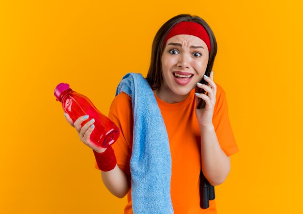 Angry young sporty woman wearing headband and wristbands with jump rope and towel on shoulders holding water bottle and talking on phone looking straight 