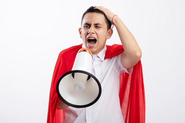 Angry young superhero boy in red cape putting hand on head looking at side shouting in loud speaker isolated on white background with copy space