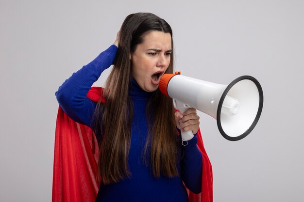 Angry young superhero girl looking down speaks on loudspeaker putting hand on head isolated on white background