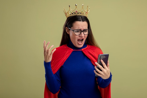 Angry young superhero girl wearing glasses and crown holding and looking at phone isolated on olive green background