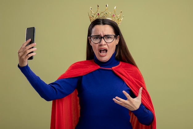 Angry young superhero girl wearing glasses and crown holding phone isolated on olive green