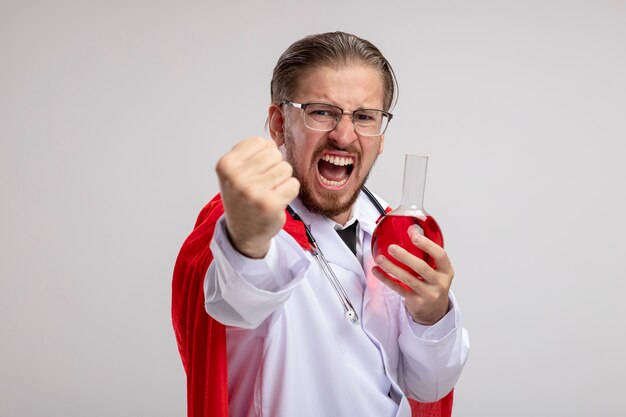 Angry young superhero guy wearing medical robe with stethoscope and glasses holding chemistry glass