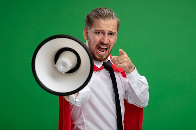Angry young superhero guy wearing tie holding out loudspeaker at camera and showing you gesture isolated on green background