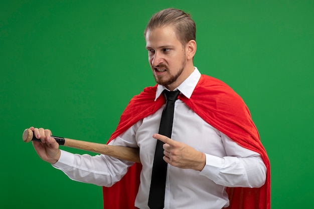 Free photo angry young superhero guy wearing tie holding and points at baseball bat isolated on green