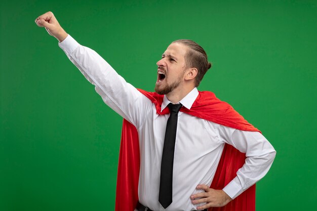 Angry young superhero guy wearing tie looking at side raising fist isolated on green background