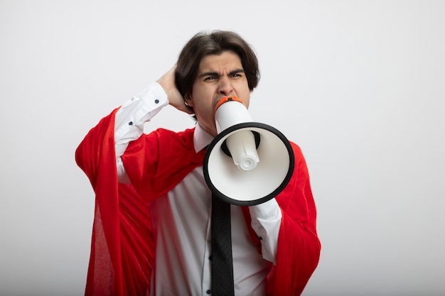 Angry young superhero guy wearing tie speaks on loudspeaker and putting hand on head isolated on white