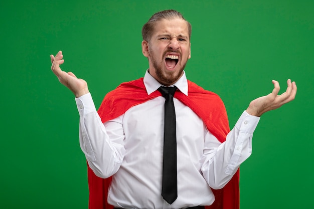 Angry young superhero guy wearing tie spreading hands isolated on green