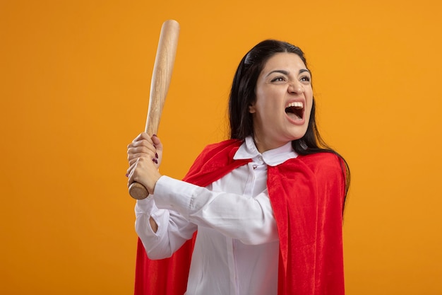 Angry young superwoman holding baseball bat looking at side isolated on orange wall