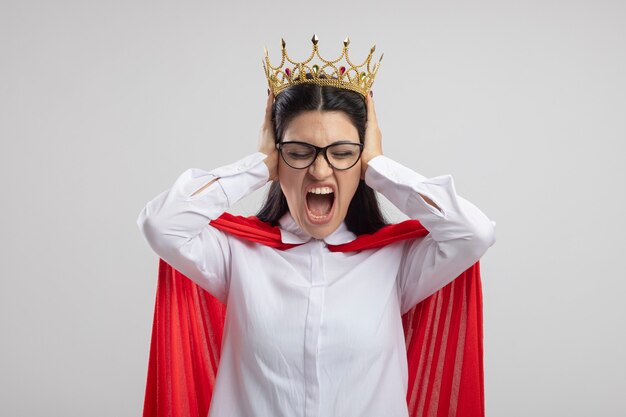 Free photo angry young superwoman wearing glasses and crown putting hands on ears screaming with closed eyes isolated on white wall