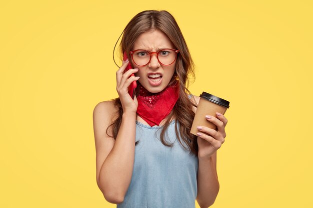 Angry young woman feels intense while has telephone conversation with friend, hears nonsense, disagrees with something, smirks face in dislike, drinks coffee, isolated over yellow wall.