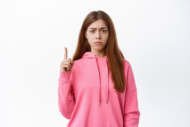 Angry young woman frowning looking with judgement and displeased face condemn something bad pointing up at logo white background