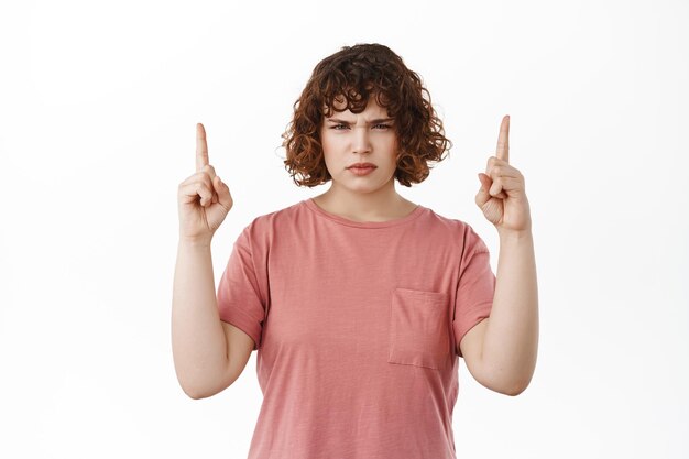 Angry young woman looks with dismay and disgust, pointing fingers up and frowning, feeling disappointed, grimacing and sulking, showing advertisement on top, white background.
