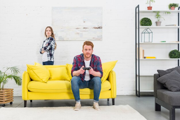 An angry young woman standing behind the yellow sofa with her boyfriend playing the video game