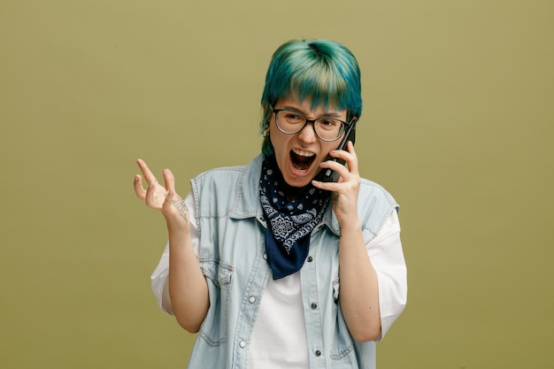 Foto gratuita giovane donna arrabbiata che indossa occhiali bandana sul collo guardando il lato che mostra la mano vuota parlando al telefono gridando isolato su sfondo verde oliva