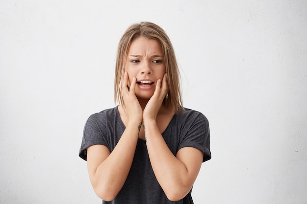 Free photo angst-ridden puzzled woman with bob hairstyle holding her hands on cheeks looking in despair. desperate fair-haired female in casual clothes having haggard look. people, emotions, feelings concept