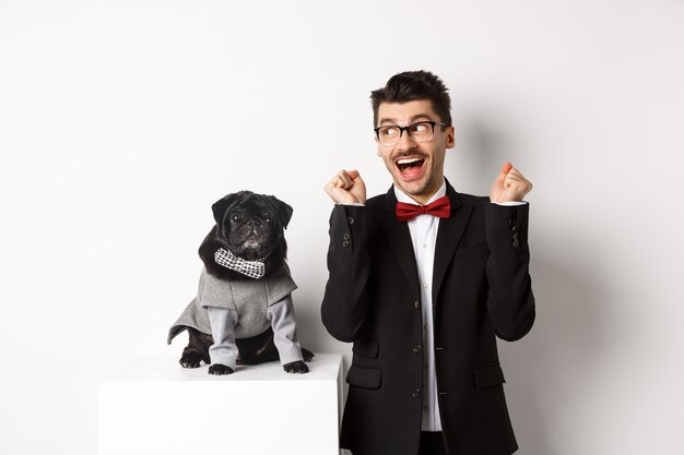 Animals, party and celebration concept. Cheerful dog owner in suit standing near cute black pug in costume, rejoicing and celebrating victory, standing over white background.