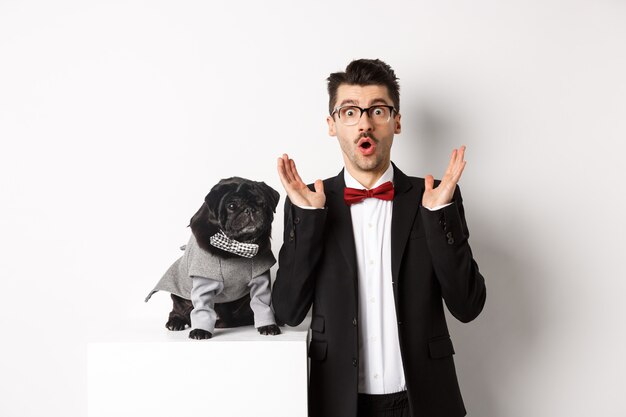 Animals, party and celebration concept. Image of dog owner and cute pug in costumes suits staring surprised at camera, reacting on promo offer, white background