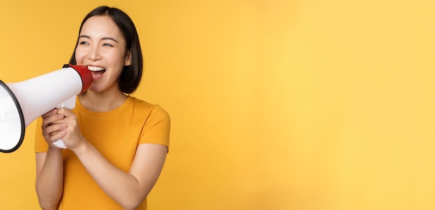 Announcement Happy asian woman shouting loud at megaphone recruiting protesting with speaker in hands standing over yellow background