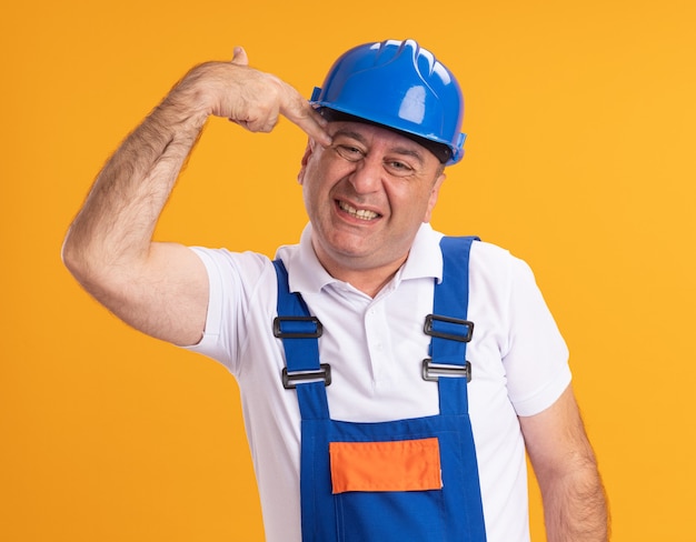 Free photo annoyed adult builder man in uniform gestures gun hand sign and holds at temple isolated on orange wall